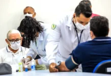 Saúde do homem: prevenção começa antes dos sintomas Homem idoso de máscara branca e óculos aguarda atendimento enquanto profissionais de saúde de jaleco realizam exames de rotina, como a aferição de pressão, em um ambiente clínico.