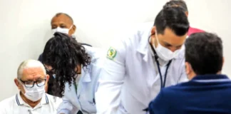 Homem idoso de máscara branca e óculos aguarda atendimento enquanto profissionais de saúde de jaleco realizam exames de rotina, como a aferição de pressão, em um ambiente clínico.