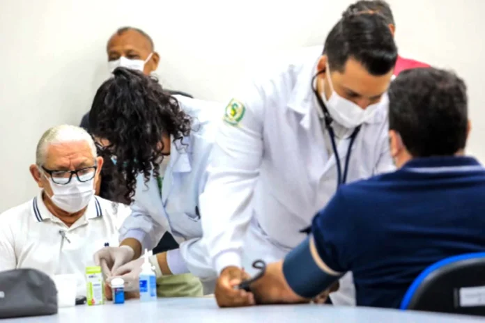 Profissionais de saúde atendem paciente homem em unidade básica de Porto Velho Homem idoso de máscara branca e óculos aguarda atendimento enquanto profissionais de saúde de jaleco realizam exames de rotina, como a aferição de pressão, em um ambiente clínico.