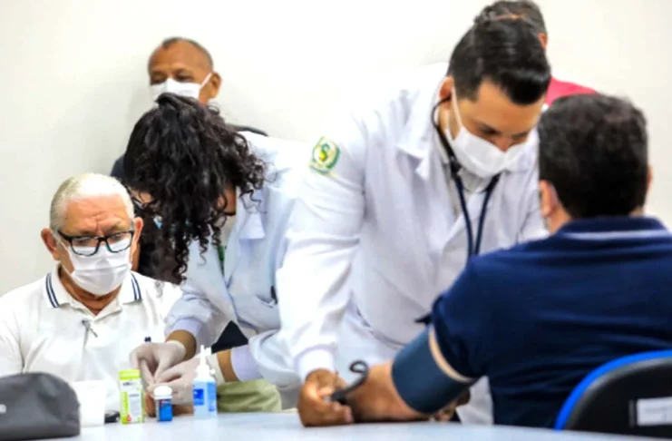 Saúde do homem: prevenção começa antes dos sintomas Homem idoso de máscara branca e óculos aguarda atendimento enquanto profissionais de saúde de jaleco realizam exames de rotina, como a aferição de pressão, em um ambiente clínico.