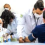 Saúde do homem: prevenção começa antes dos sintomas Homem idoso de máscara branca e óculos aguarda atendimento enquanto profissionais de saúde de jaleco realizam exames de rotina, como a aferição de pressão, em um ambiente clínico.