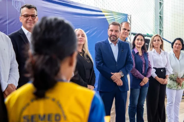 Hildon Chaves em evento da educação com profissionais da Semed O candidato Hildon Chaves, vestindo terno azul, sorri enquanto está rodeado por profissionais da educação e autoridades durante um evento em uma quadra escolar.