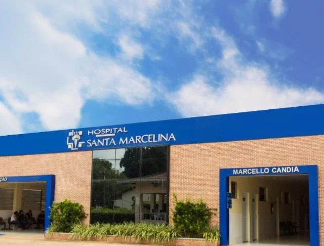 Hospital Santa Marcelina realiza 2ª Feijoada Solidária Fotografia da fachada do Hospital Santa Marcelina, destacando o prédio de tijolos aparentes com letreiro azul e branco e a entrada do setor Marcello Candia, sob um céu claro com nuvens.