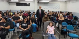 MPRO debate consumo consciente com acadêmicos de Direito O promotor de Justiça Alan Castiel Barbosa, de terno e gravata, posa sorridente em pé no fundo de uma sala de aula universitária, acompanhado por dezenas de estudantes de Direito acomodados em cadeiras azuis.