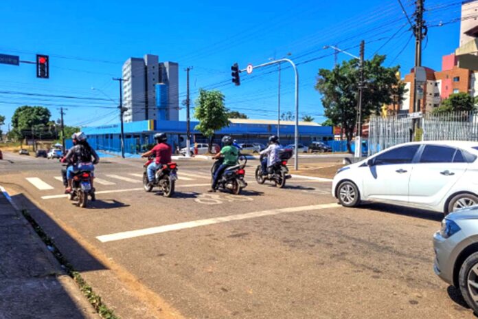 Motociclistas aguardam no semáforo em cruzamento de Porto Velho Trânsito em Porto Velho, mostrando vários motociclistas e um carro branco parados aguardando o semáforo abrir em um dia ensolarado.