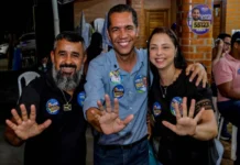 O candidato ao Senado, Luis Fernando, veste camisa azul e sorri para a foto ao lado de dois apoiadores, todos com as mãos abertas indicando o número 55.