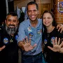 O candidato ao Senado, Luis Fernando, veste camisa azul e sorri para a foto ao lado de dois apoiadores, todos com as mãos abertas indicando o número 55.
