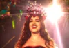 A cantora Anitta sorridente e iluminada, a usar uma coroa vermelha cravejada de pedras e búzios durante o evento da Grande Rio.