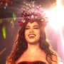 A cantora Anitta sorridente e iluminada, a usar uma coroa vermelha cravejada de pedras e búzios durante o evento da Grande Rio.