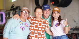 Mariana Carvalho, usando boné e camisa de campanha, sorri e abraça calorosamente três apoiadores durante um evento eleitoral no interior de Rondônia.