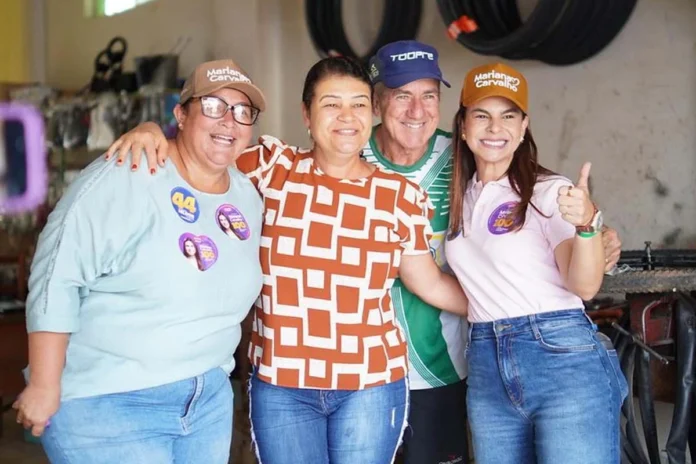 Mariana Carvalho, usando boné e camisa de campanha, sorri e abraça calorosamente três apoiadores durante um evento eleitoral no interior de Rondônia.