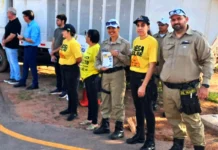 Agentes de trânsito e outros participantes, alguns vestindo camisetas amarelas, estão posicionados em uma rua durante um ato cívico em Rolim de Moura.
