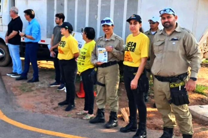 Agentes de trânsito e outros participantes, alguns vestindo camisetas amarelas, estão posicionados em uma rua durante um ato cívico em Rolim de Moura.