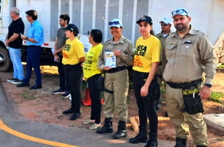 Agentes de trânsito e outros participantes, alguns vestindo camisetas amarelas, estão posicionados em uma rua durante um ato cívico em Rolim de Moura.