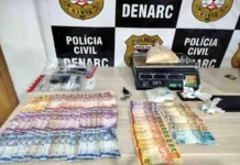Denarc prende dupla e apreende 1kg de cocaína na Zona Sul Mesa com materiais apreendidos pela polícia, incluindo cédulas de dinheiro espalhadas, uma balança de precisão digital com um saco de substância branca e outros invólucros menores, em frente a um banner do DENARC - Polícia Civil.