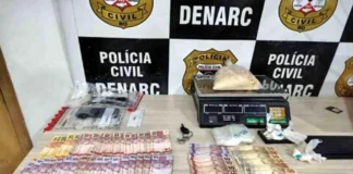 Denarc prende dupla e apreende 1kg de cocaína na Zona Sul Mesa com materiais apreendidos pela polícia, incluindo cédulas de dinheiro espalhadas, uma balança de precisão digital com um saco de substância branca e outros invólucros menores, em frente a um banner do DENARC - Polícia Civil.