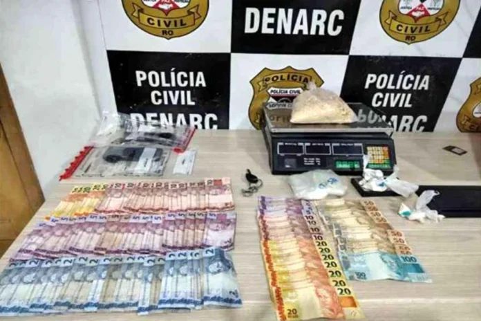 Mesa com materiais apreendidos pela polícia, incluindo cédulas de dinheiro espalhadas, uma balança de precisão digital com um saco de substância branca e outros invólucros menores, em frente a um banner do DENARC - Polícia Civil.