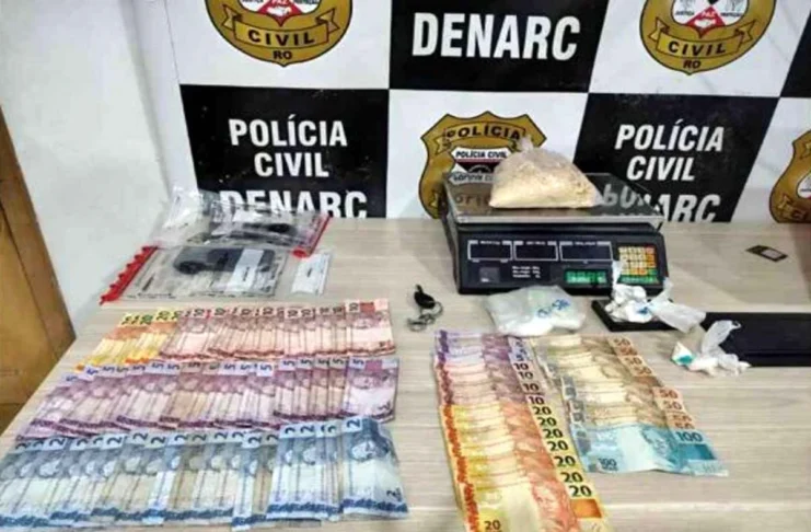 Denarc prende dupla e apreende 1kg de cocaína na Zona Sul Mesa com materiais apreendidos pela polícia, incluindo cédulas de dinheiro espalhadas, uma balança de precisão digital com um saco de substância branca e outros invólucros menores, em frente a um banner do DENARC - Polícia Civil.