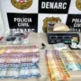 Denarc prende dupla e apreende 1kg de cocaína na Zona Sul Mesa com materiais apreendidos pela polícia, incluindo cédulas de dinheiro espalhadas, uma balança de precisão digital com um saco de substância branca e outros invólucros menores, em frente a um banner do DENARC - Polícia Civil.