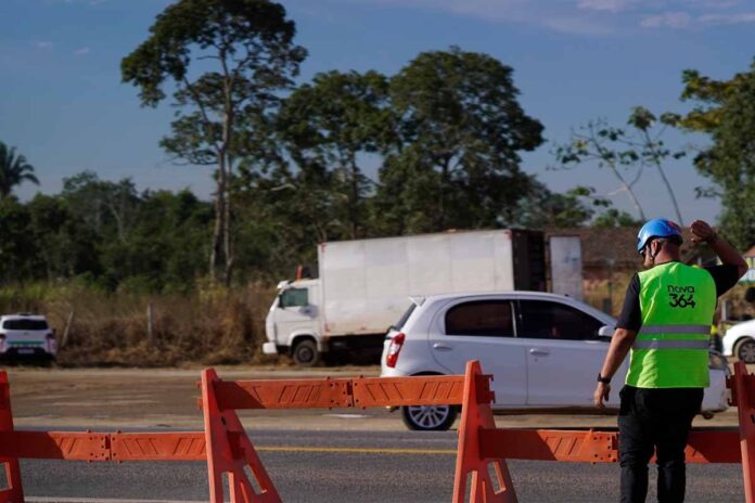 Fiscalização e obras na rodovia Nova 364 Funcionário com colete da Nova 364 a sinalizar a rodovia junto a barreiras de segurança e veículos em trânsito.