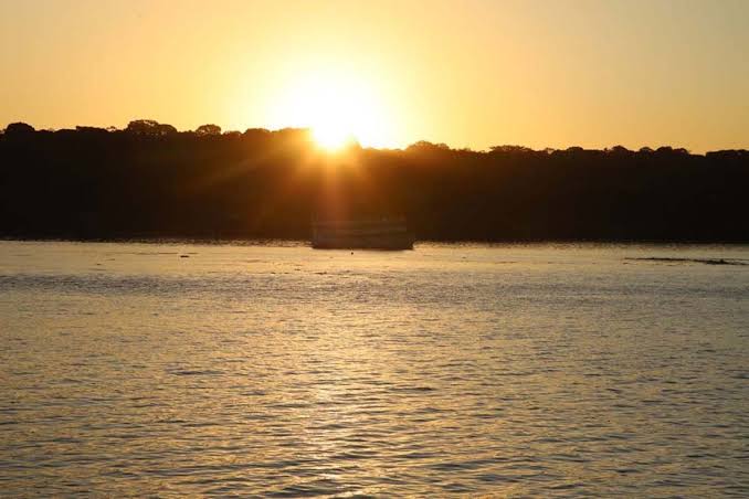 Pôr do sol com calor intenso em Rondônia Fotografia de um pôr do sol alaranjado sobre um rio, ilustrando o clima quente da região.