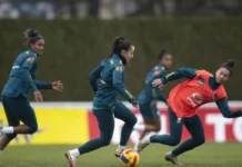Jogadoras da seleção brasileira feminina durante um treinamento no gramado, vestindo uniformes azuis e laranjas com o escudo da CBF.
