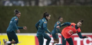 Jogadoras da seleção brasileira feminina durante um treinamento no gramado, vestindo uniformes azuis e laranjas com o escudo da CBF.