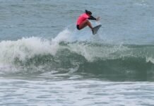 Inscrições abertas para o 30º Circuito A Tribuna de Surf Colegial no Guarujá Uma surfista competidora, vestindo uma lycra rosa e parte de baixo preta, realiza uma manobra aérea sobre as ondas durante a bateria de surfe.
