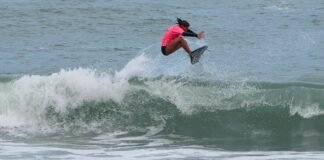 Inscrições abertas para o 30º Circuito A Tribuna de Surf Colegial no Guarujá Uma surfista competidora, vestindo uma lycra rosa e parte de baixo preta, realiza uma manobra aérea sobre as ondas durante a bateria de surfe.