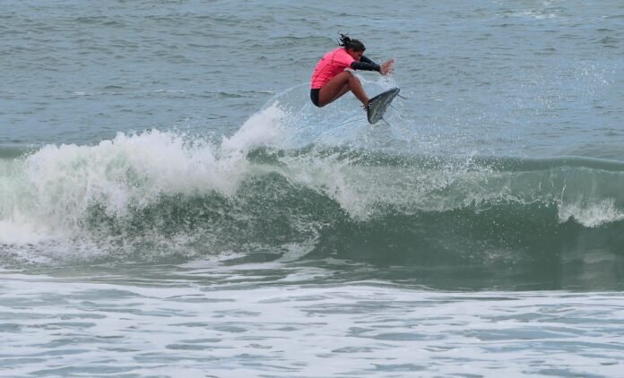 Surfista em ação no Circuito A Tribuna de Surf Colegial Uma surfista competidora, vestindo uma lycra rosa e parte de baixo preta, realiza uma manobra aérea sobre as ondas durante a bateria de surfe.