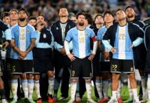 O jogador Lionel Messi em pé no gramado do estádio, olhando para o alto com uma expressão de claro desconforto e frustração.