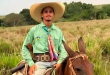 Fotografia do jovem Marcos André Ferreira Santos montado a cavalo, sorrindo e vestindo um chapéu de palha, camisa verde e lenço colorido no pescoço em uma área de pastagem.