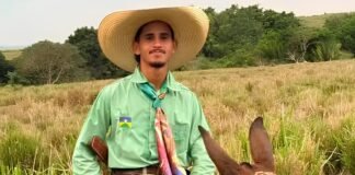 Fotografia do jovem Marcos André Ferreira Santos montado a cavalo, sorrindo e vestindo um chapéu de palha, camisa verde e lenço colorido no pescoço em uma área de pastagem.