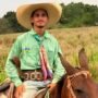 Fotografia do jovem Marcos André Ferreira Santos montado a cavalo, sorrindo e vestindo um chapéu de palha, camisa verde e lenço colorido no pescoço em uma área de pastagem.