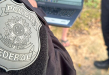 PF investiga policiais ligados à facção no RJ Primeiro plano do brasão da Polícia Federal bordado no uniforme preto de um agente. Ao fundo, de forma desfocada, há um laptop sendo utilizado ao ar livre e pessoas acompanhando a ação no local.
