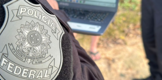 PF investiga policiais ligados à facção no RJ Primeiro plano do brasão da Polícia Federal bordado no uniforme preto de um agente. Ao fundo, de forma desfocada, há um laptop sendo utilizado ao ar livre e pessoas acompanhando a ação no local.