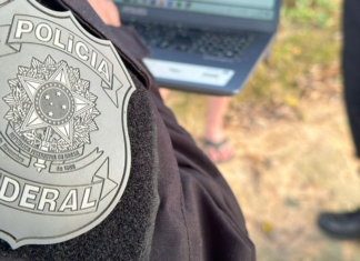 PF investiga policiais ligados à facção no RJ Primeiro plano do brasão da Polícia Federal bordado no uniforme preto de um agente. Ao fundo, de forma desfocada, há um laptop sendo utilizado ao ar livre e pessoas acompanhando a ação no local.