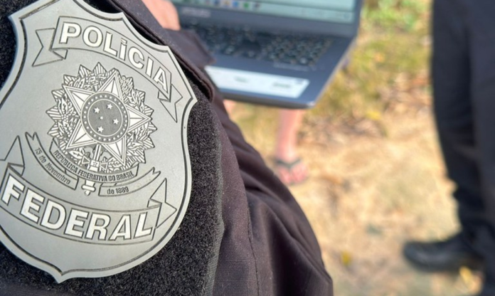 Primeiro plano do brasão da Polícia Federal bordado no uniforme preto de um agente. Ao fundo, de forma desfocada, há um laptop sendo utilizado ao ar livre e pessoas acompanhando a ação no local.