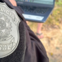 PF investiga policiais ligados à facção no RJ Primeiro plano do brasão da Polícia Federal bordado no uniforme preto de um agente. Ao fundo, de forma desfocada, há um laptop sendo utilizado ao ar livre e pessoas acompanhando a ação no local.