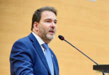 O deputado estadual Alex Redano a discursar num púlpito ao microfone, envergando um fato azul-escuro e gravata azul-clara.