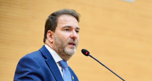 O deputado estadual Alex Redano a discursar num púlpito ao microfone, envergando um fato azul-escuro e gravata azul-clara.