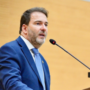 O deputado estadual Alex Redano a discursar num púlpito ao microfone, envergando um fato azul-escuro e gravata azul-clara.