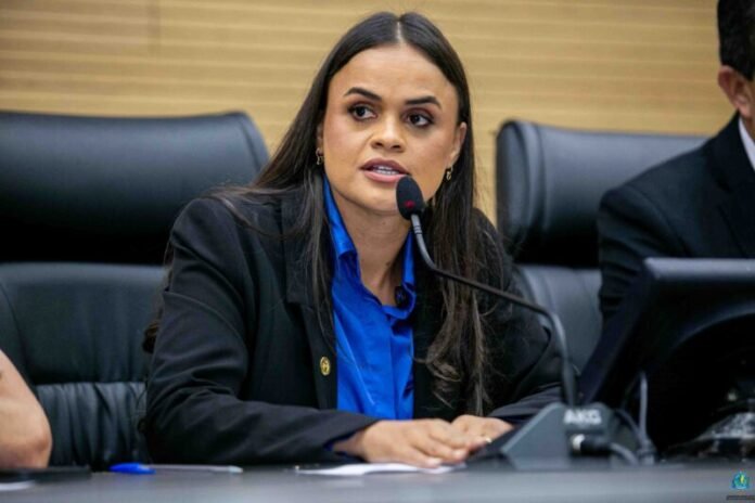 Deputada Dra Taissa Sousa discursa no parlamento A deputada estadual Dra. Taíssa Sousa a discursar ao microfone durante uma sessão parlamentar, envergando um casaco escuro e uma camisa azul.