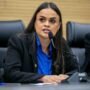 A deputada estadual Dra. Taíssa Sousa a discursar ao microfone durante uma sessão parlamentar, envergando um casaco escuro e uma camisa azul.