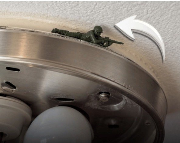 Soldadinho escondido na luminária do teto Imagem aproxima um pequeno soldadinho de plástico verde posicionado deitado, de barriga para baixo, sobre a borda metálica de uma luminária de teto, apontando sua arma para fora.