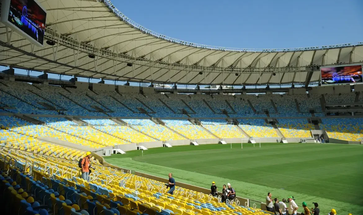 Vista interior das bancadas vazias pintadas de azul e amarelo e do relvado verde do Estádio do Maracanã, no Rio de Janeiro, num dia de sol.