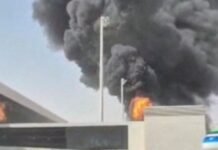 Densa nuvem de fumaça escura e chamas intensas subindo por trás do muro e da estrutura do terminal de um aeroporto, evidenciando uma forte explosão.