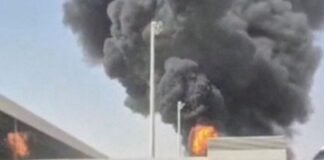 Após ataque fatal, houthis ameaçam espaço aéreo e paralisam voos na Arábia Saudita Densa nuvem de fumaça escura e chamas intensas subindo por trás do muro e da estrutura do terminal de um aeroporto, evidenciando uma forte explosão.