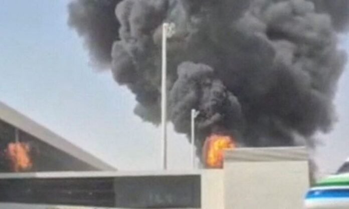Densa nuvem de fumaça escura e chamas intensas subindo por trás do muro e da estrutura do terminal de um aeroporto, evidenciando uma forte explosão.