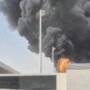 Densa nuvem de fumaça escura e chamas intensas subindo por trás do muro e da estrutura do terminal de um aeroporto, evidenciando uma forte explosão.